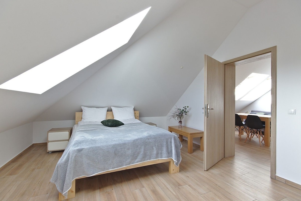 Un dormitorio con techo inclinado, una cama, mesitas de noche y vista hacia un comedor.