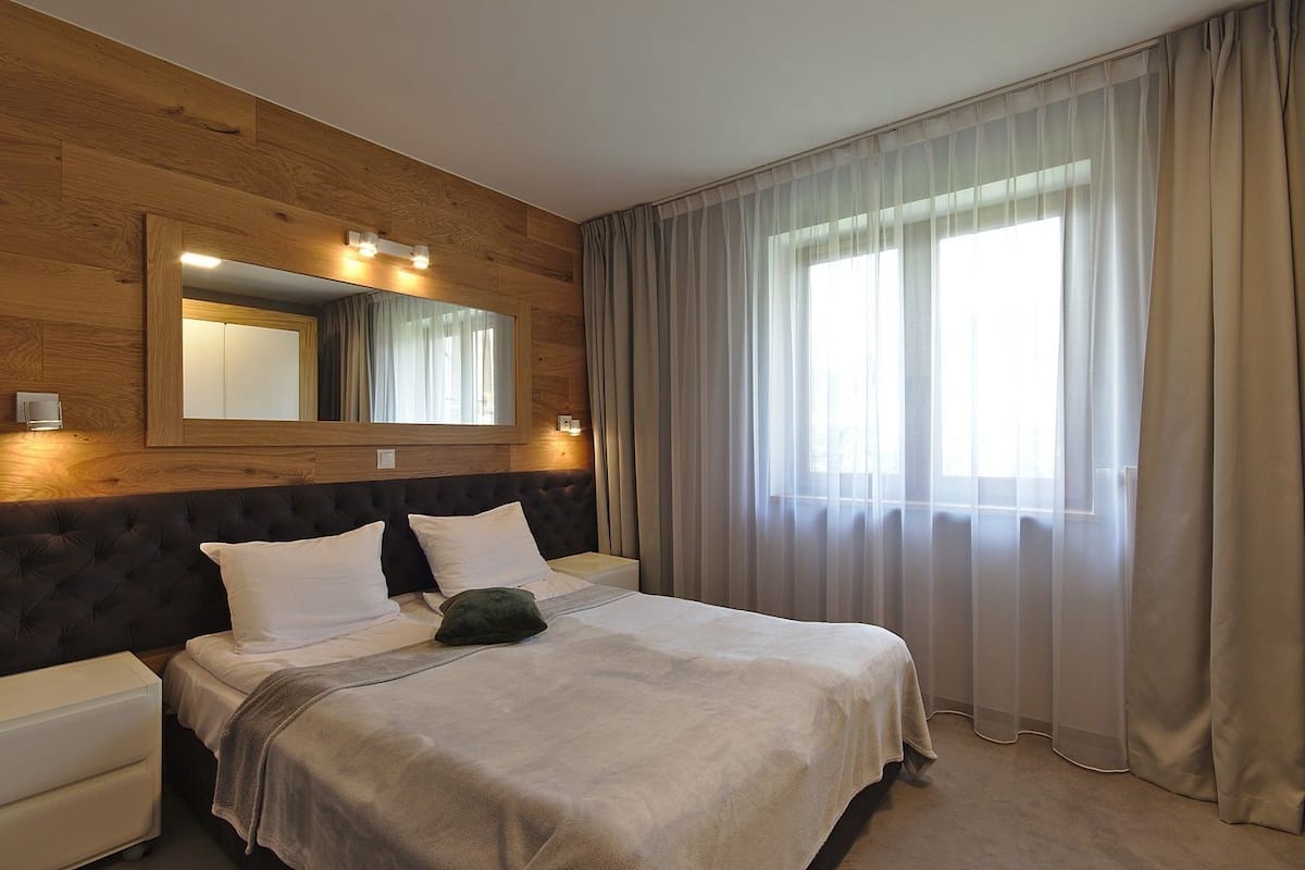 Habitación de hotel con cabecera de madera, un espejo grande, una cama con sábanas blancas, un cojín verde y cortinas de gasa.