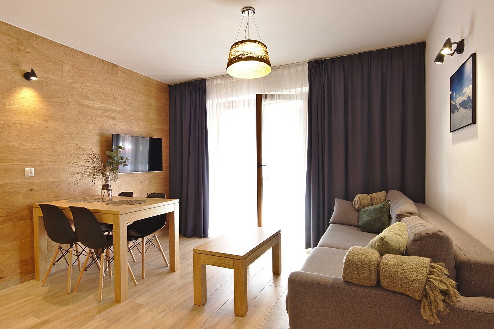 Un salon moderne avec un mur en bois, un canapé avec des coussins, une table à manger avec des chaises, un téléviseur fixé au mur et une grande fenêtre avec des rideaux.