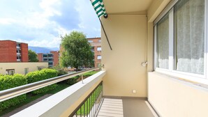 A balcony with a metal railing, a window with a floral curtain, and a view of a tree and buildings in the background. - Girasole (Savosa)
