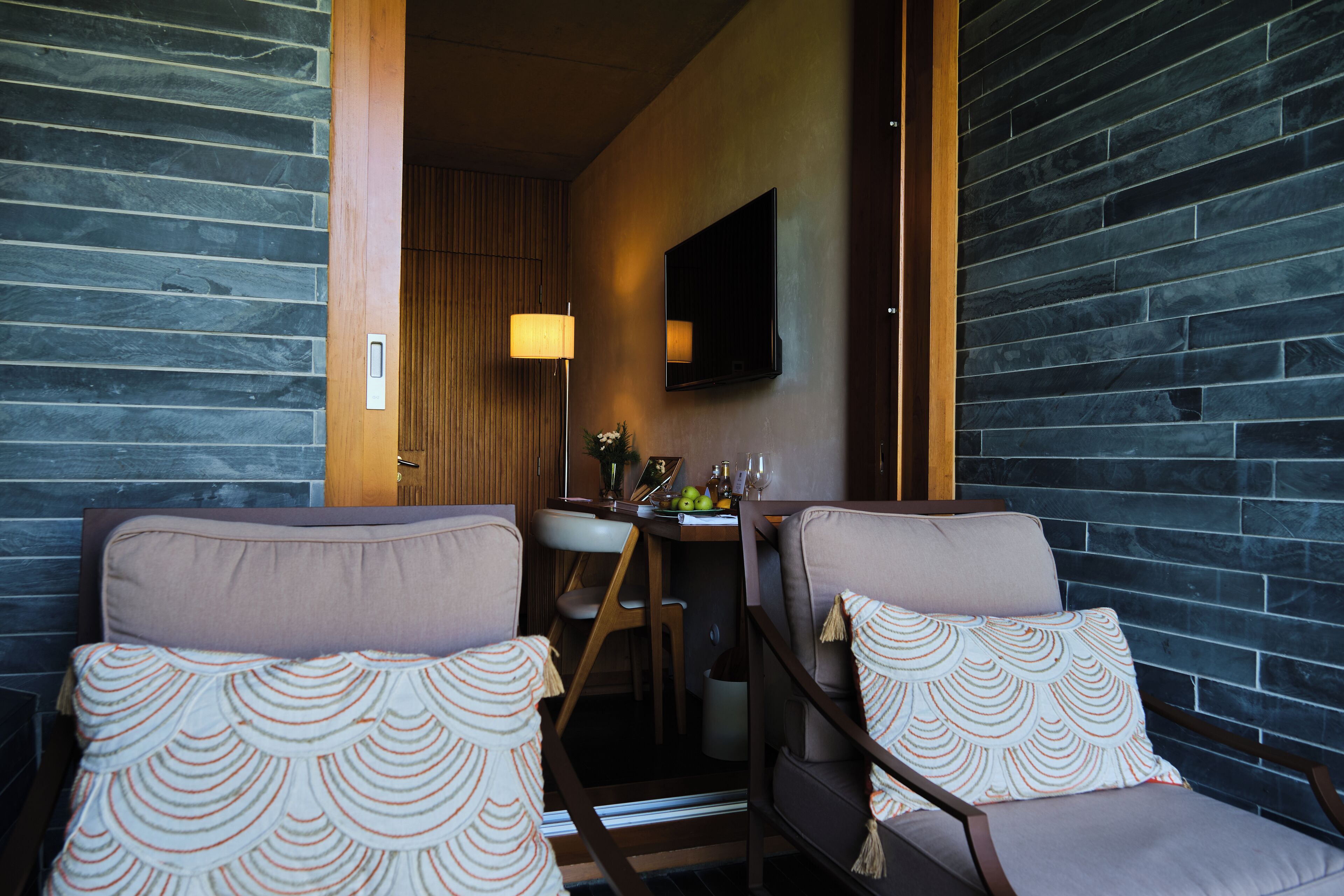 A room with two cushioned chairs, a wooden table, a flat-screen TV, and a stone wall.