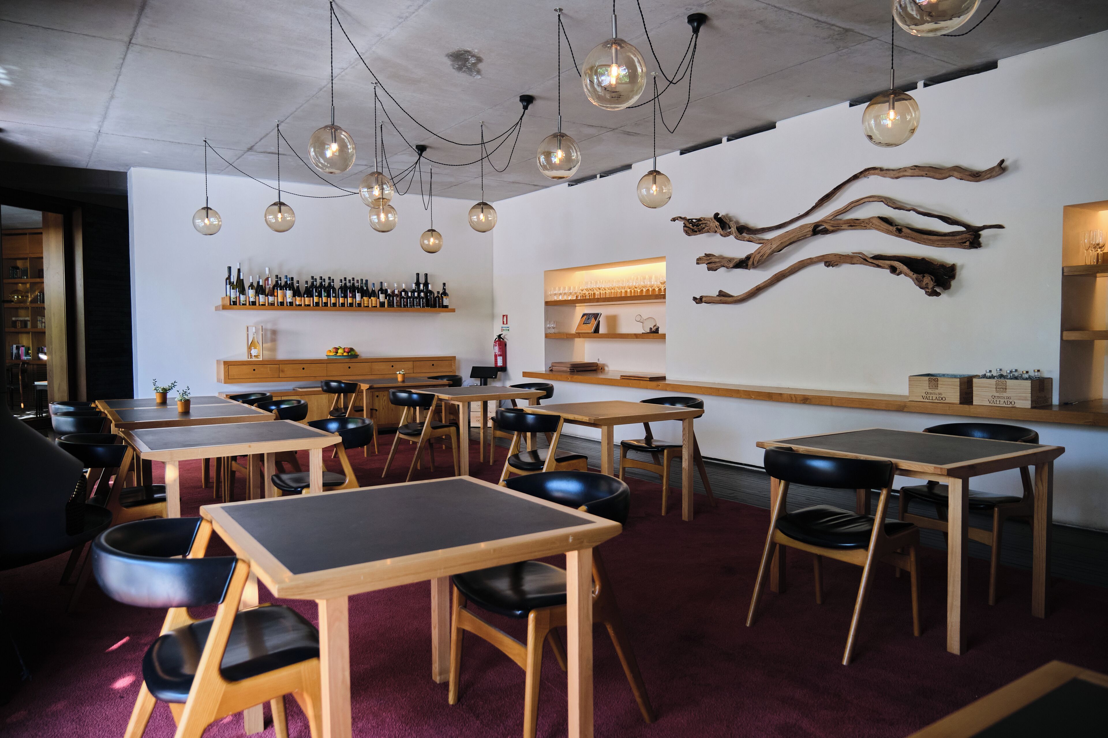 A restaurant interior with wooden tables and black chairs, a bar area with bottles and glasses, and a wall with wooden branches.