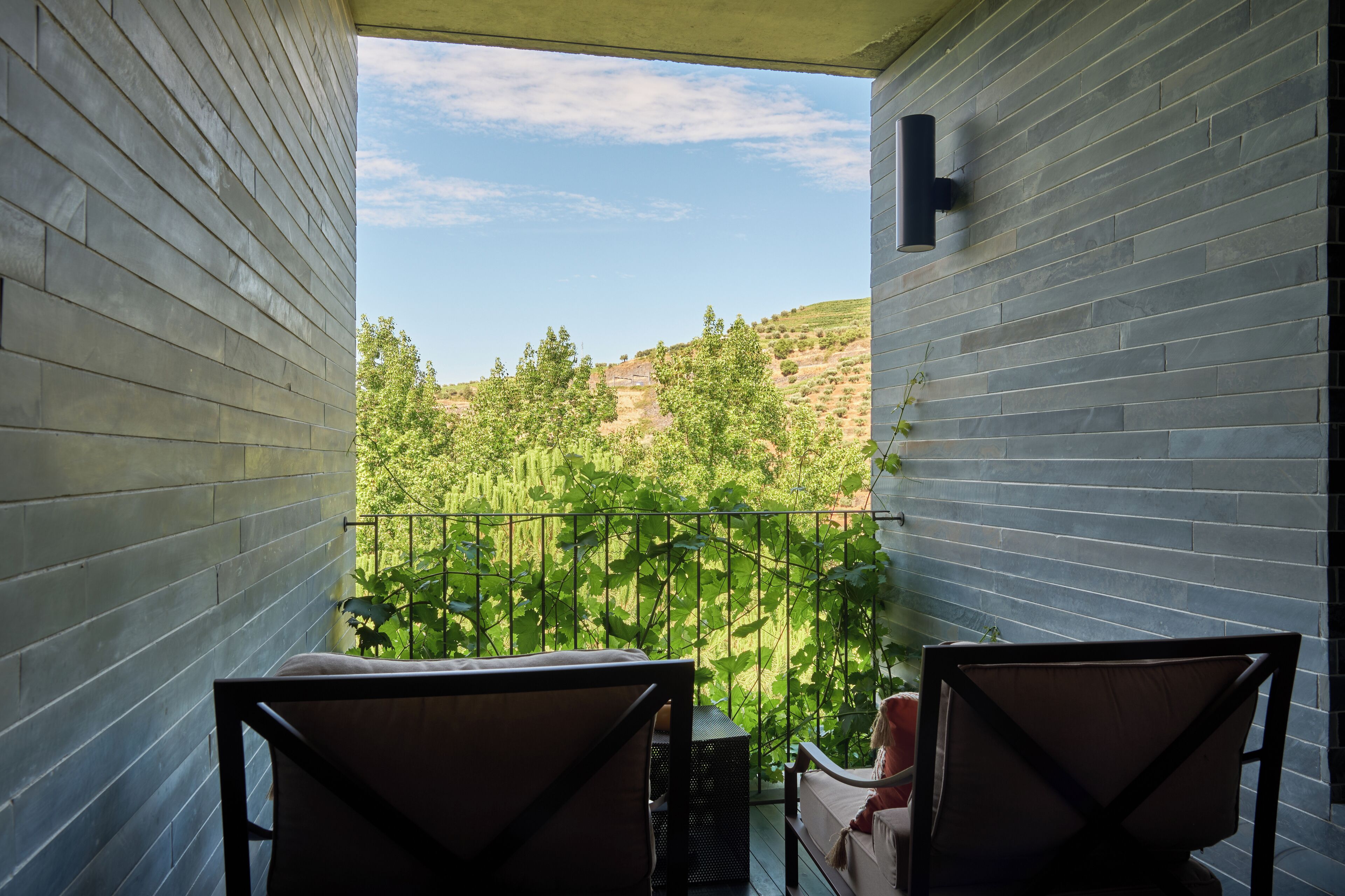 Un balcón con dos sillas, una mesa pequeña y vistas a un paisaje verde con árboles y colinas.