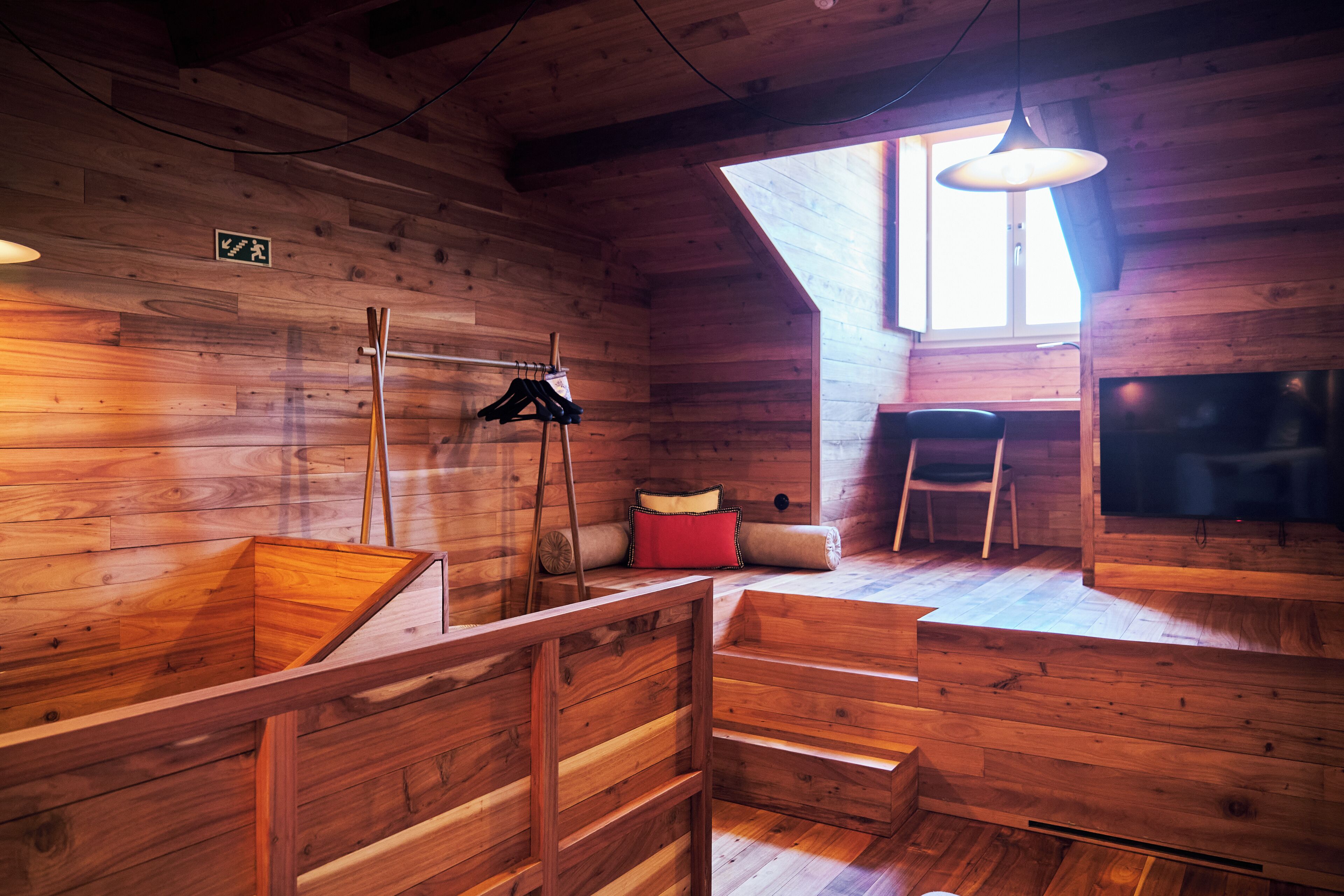 Interior de una cabaña de madera con una cama en el altillo, una silla, una lámpara colgante y una chimenea.