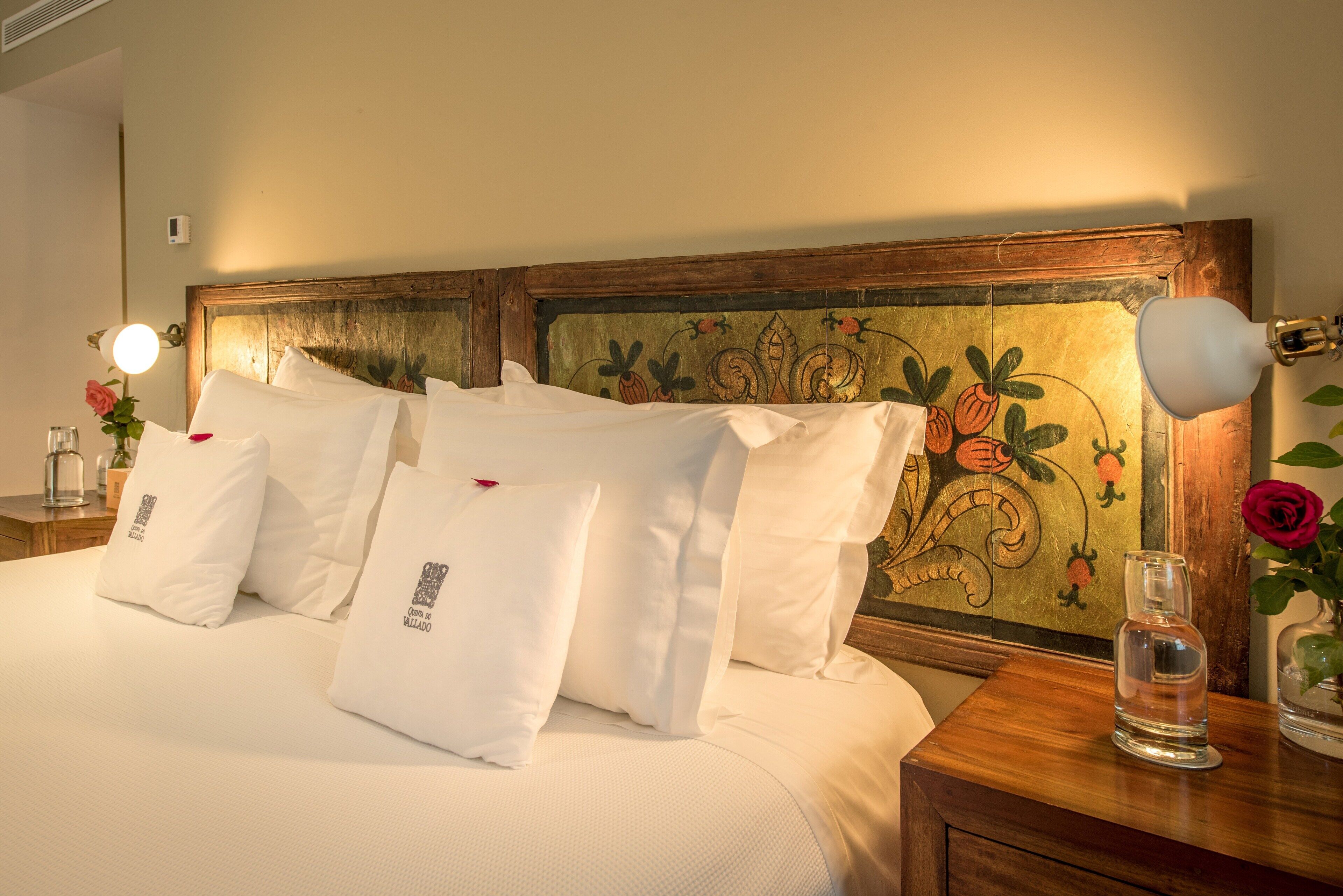 Una cama bien tendida con sábanas blancas y varias almohadas, un cabecero de madera con detalles florales, una mesita de noche con una lámpara y un jarrón con una rosa.