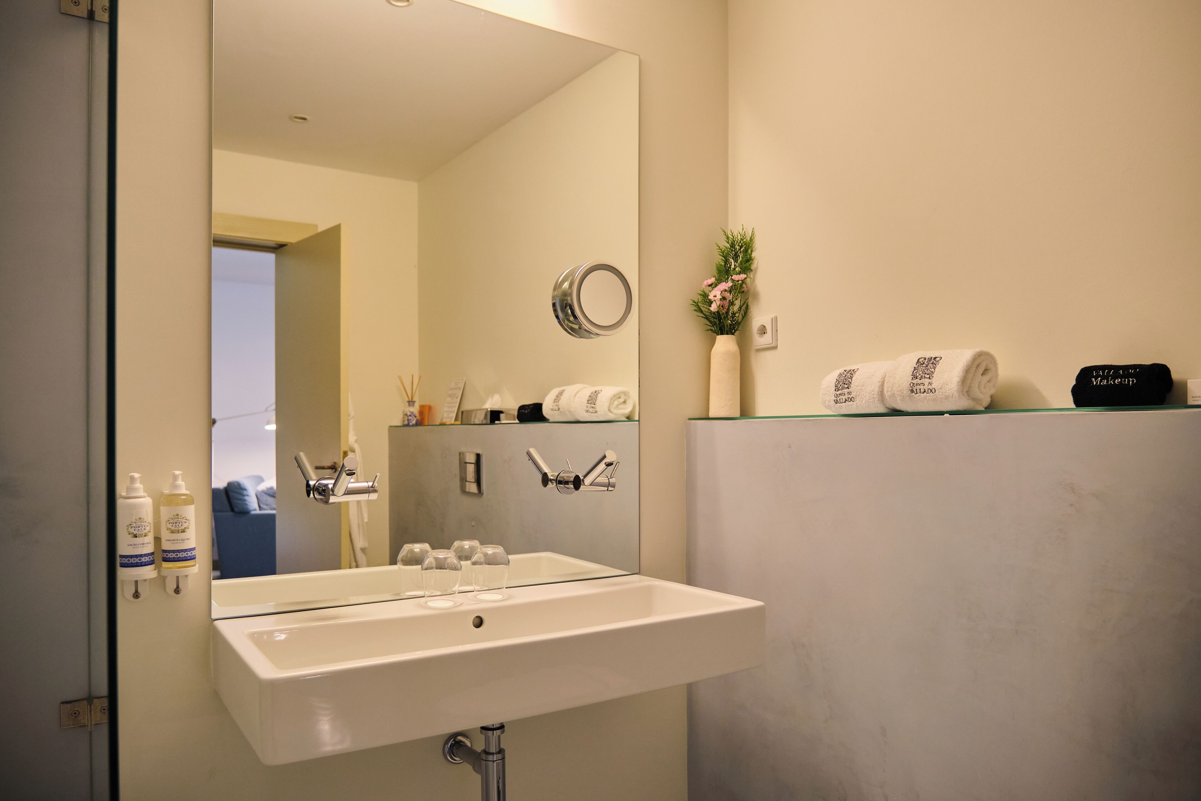 Un baño con un lavamanos blanco, un espejo, un perchero para toallas y un estante con toallas y un artículo de maquillaje.