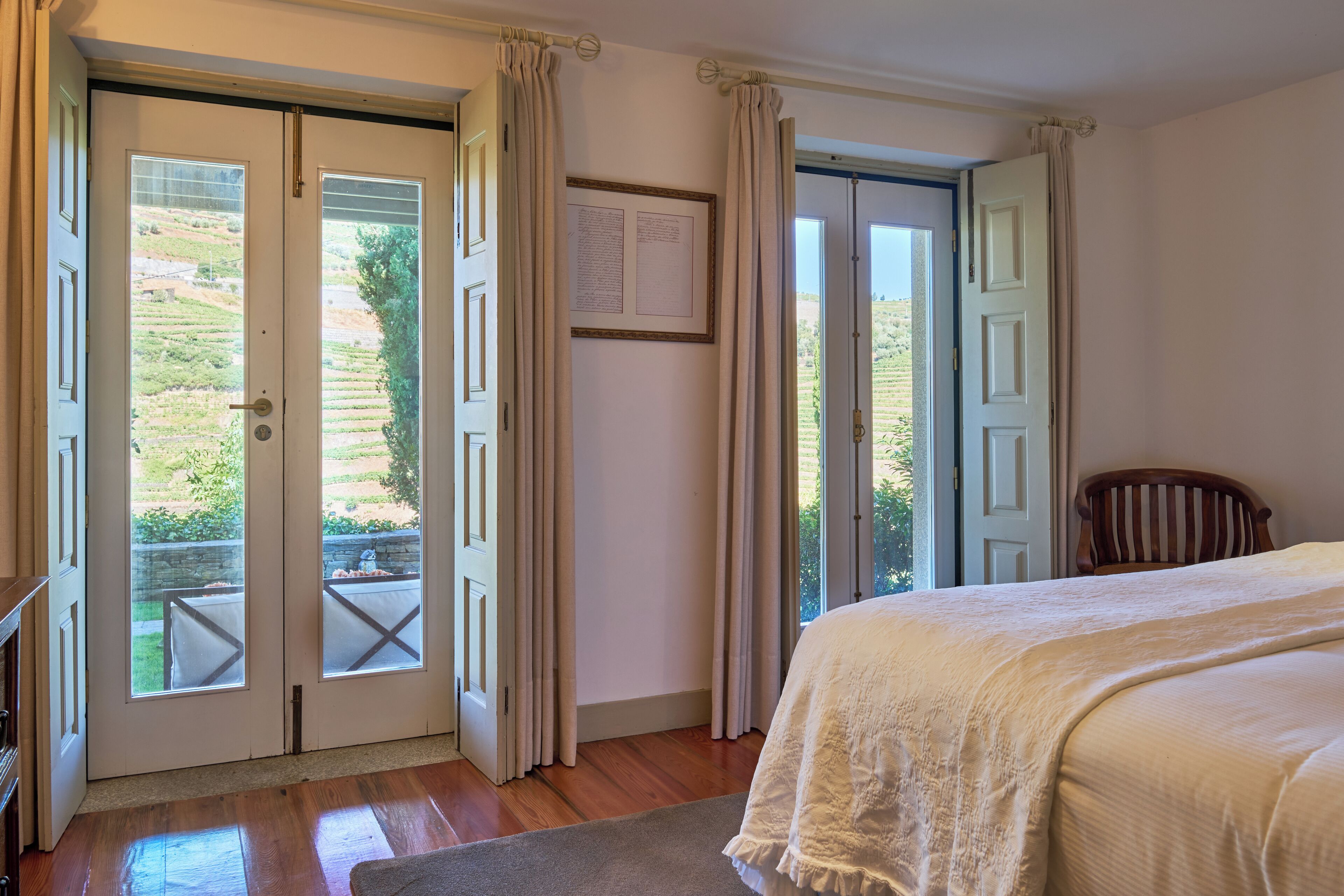 A bedroom with a large bed, wooden flooring, and a view through a door to an outdoor area with a table and chairs.