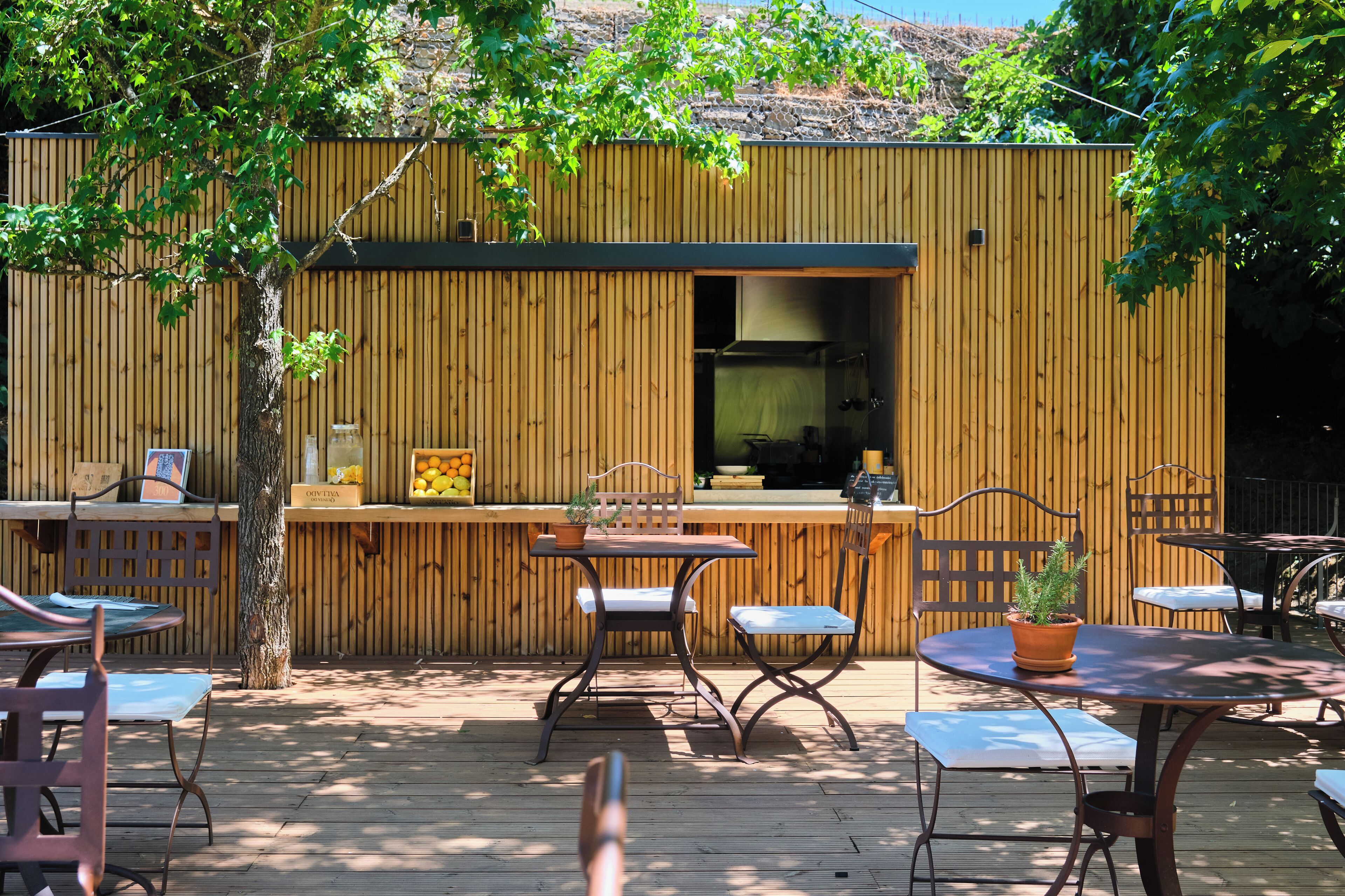 Eine hölzerne Terrasse im Freien mit Tischen und Stühlen, ein Barbereich mit einem Blumentopf und ein Holzzaun im Hintergrund.