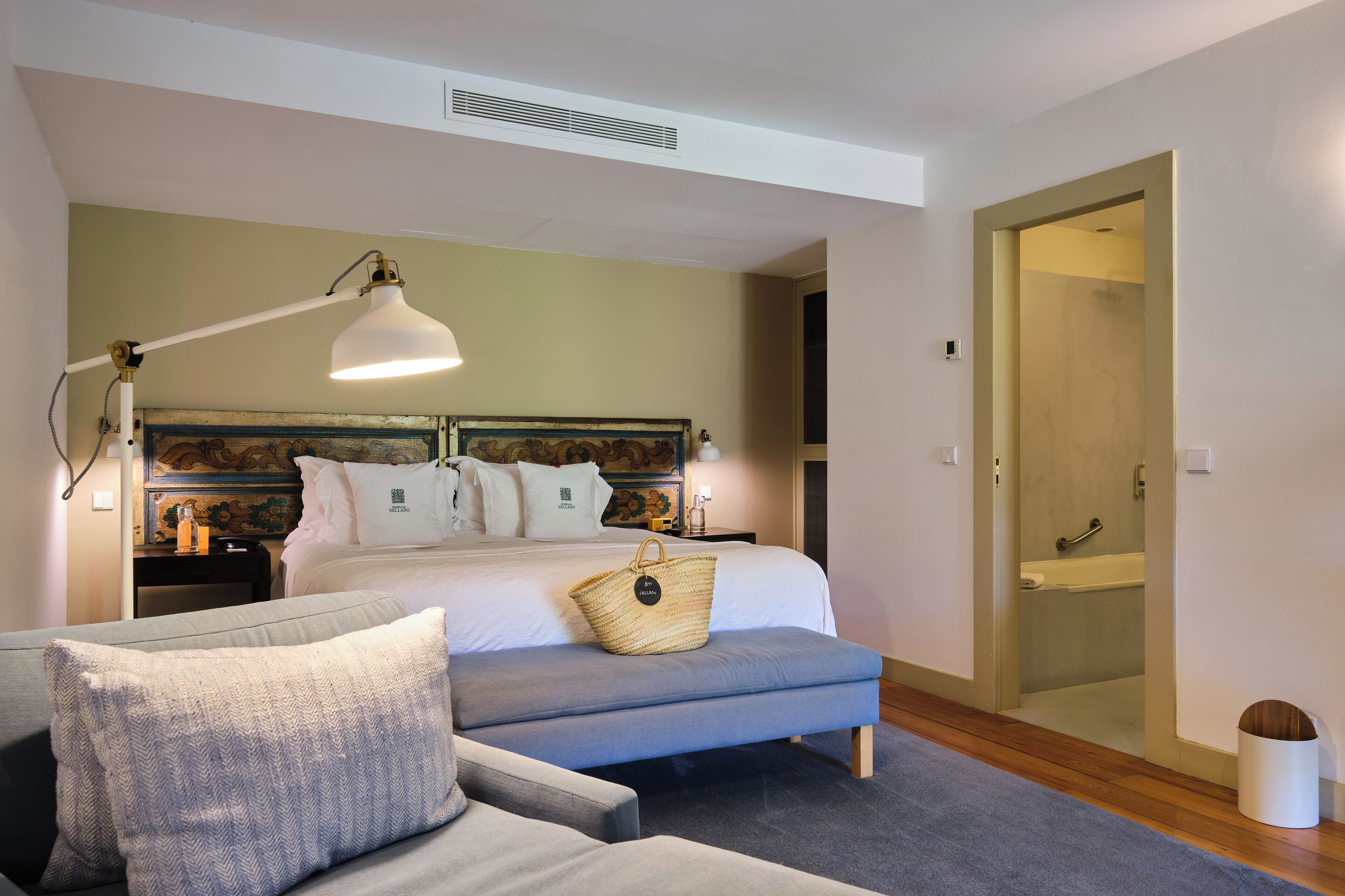 A bedroom with a bed, bedside tables, a lamp, a bench, and a bathroom visible through an open door.