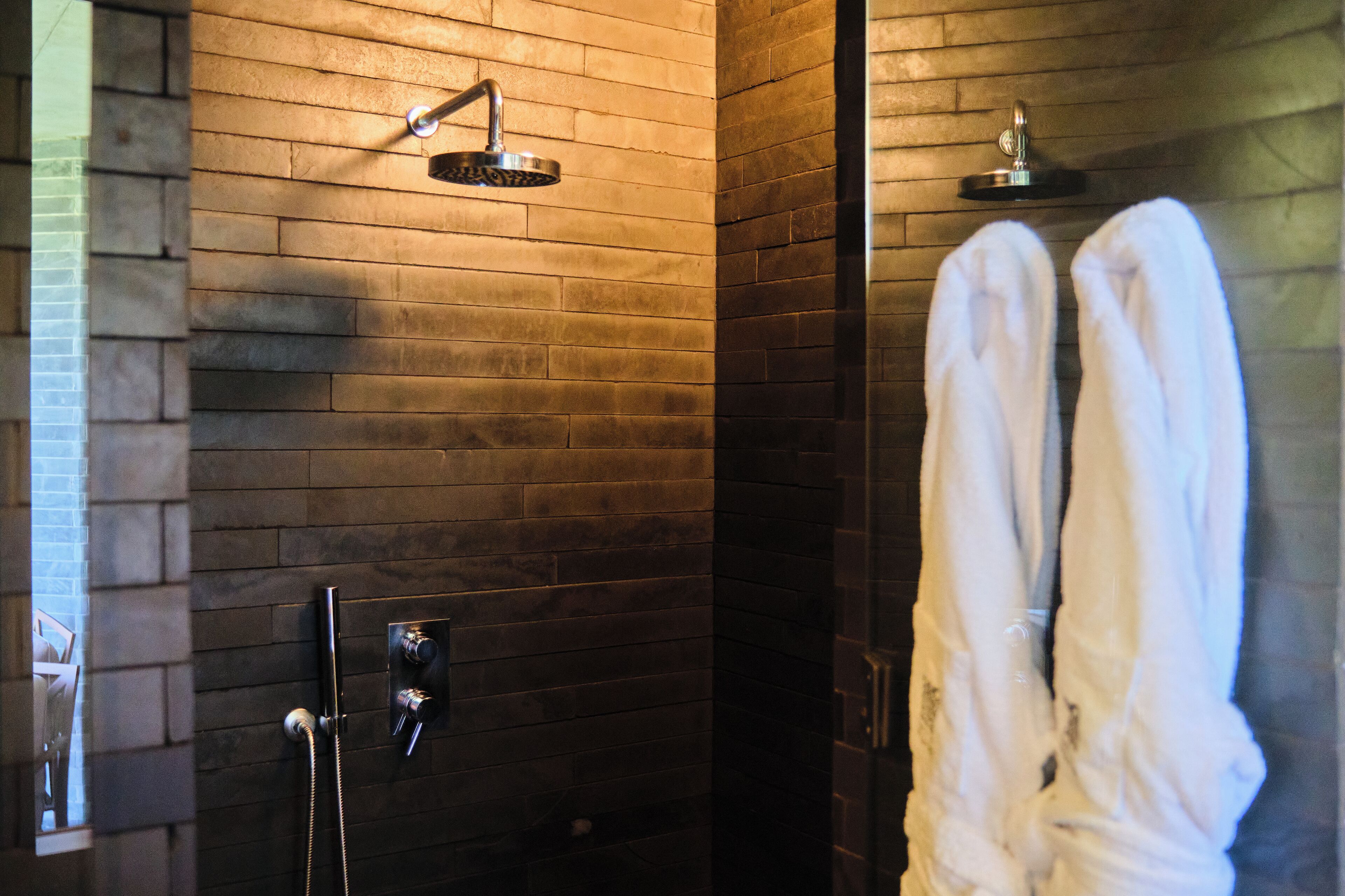 Une salle de bain moderne avec un mur en briques, une douche équipée d’un pommeau de douche à main et deux serviettes blanches suspendues à des crochets.