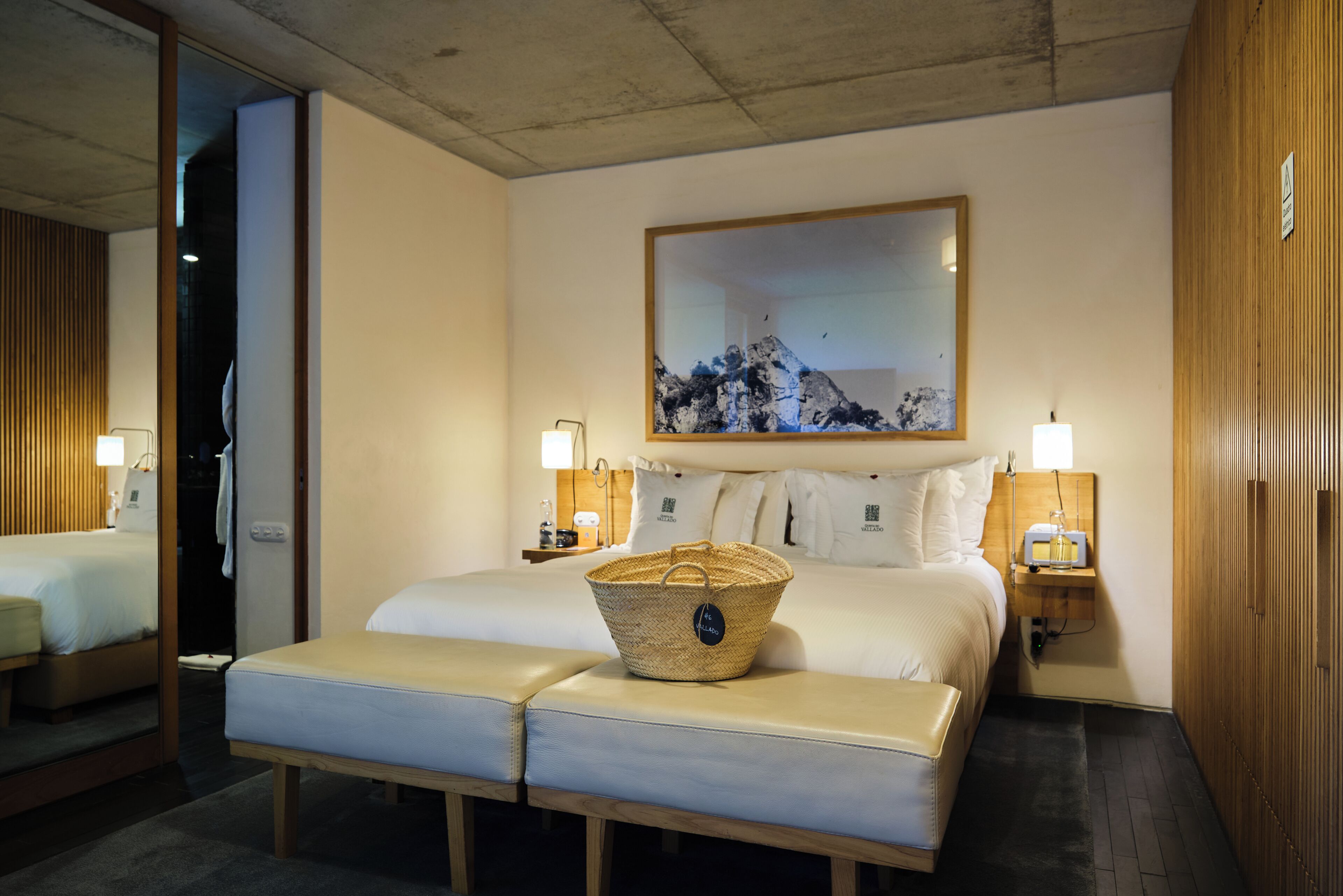 Quarto de hotel com uma cama grande, duas mesas de cabeceira com abajures, um banco de madeira e uma grande obra de arte emoldurada na parede.