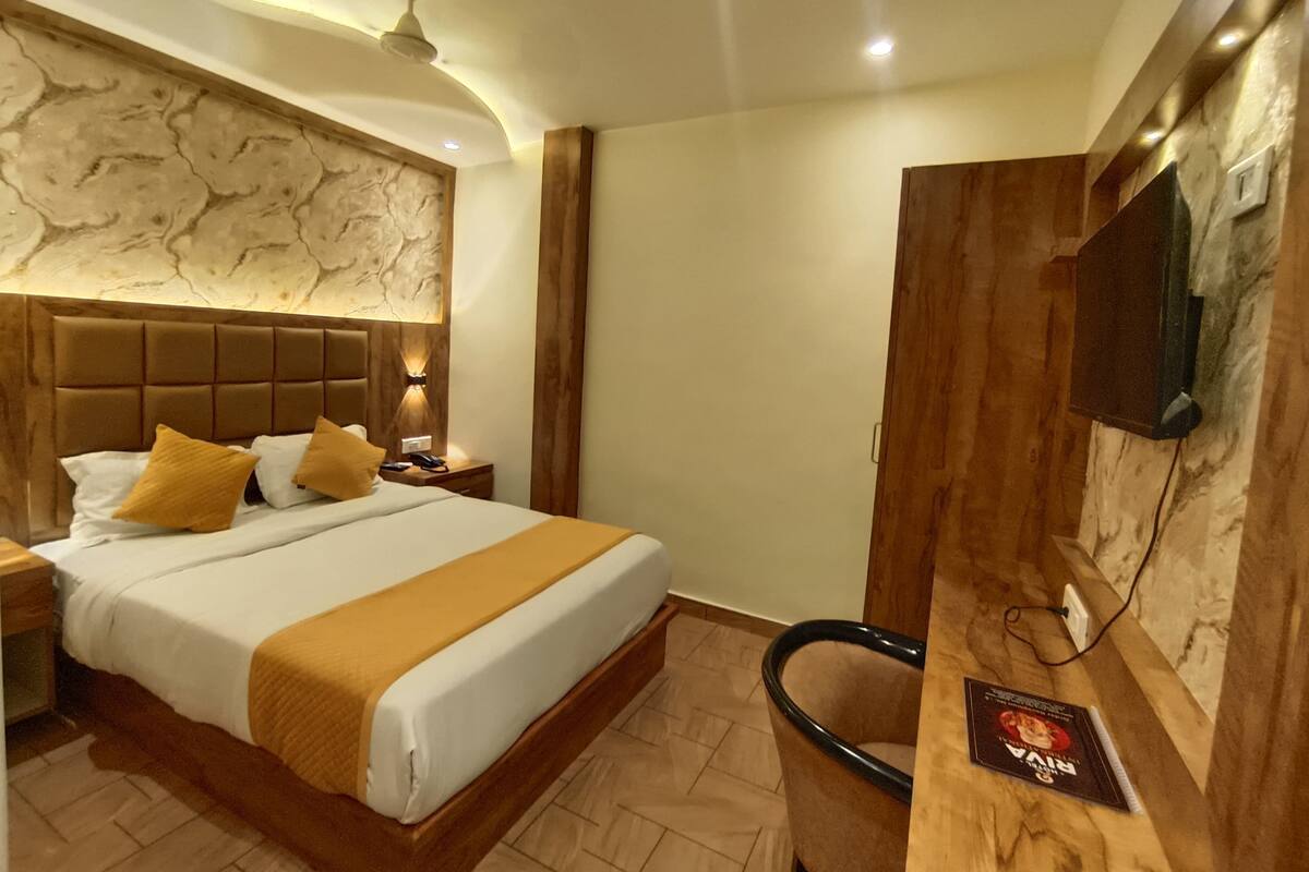 A hotel room with a large bed, a wooden headboard, a desk with a chair, a flat-screen TV mounted on the wall, and a ceiling fan.