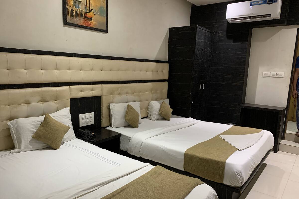 A hotel room with two beds, a large painting on the wall, a black wardrobe, and an air conditioning unit.