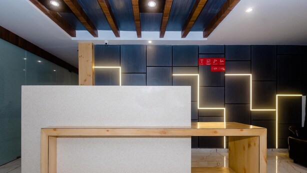 A reception desk with a wooden frame and a white top, set against a wall with geometric light patterns and a red sign with icons. - Hotel Riva International - Near Nesco Center (Mumbai)