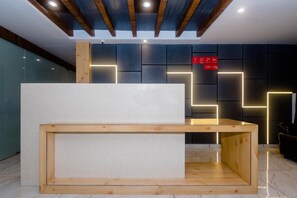 A reception desk with a wooden frame and a white top, set against a wall with geometric light patterns and a red sign with icons. - Hotel Riva International - Near Nesco Center (Mumbai)