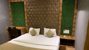 A bedroom with a tufted headboard, a wooden desk, a chair, and a bedside table with a power outlet. - Hotel Riva International - Near Nesco Center (Mumbai)