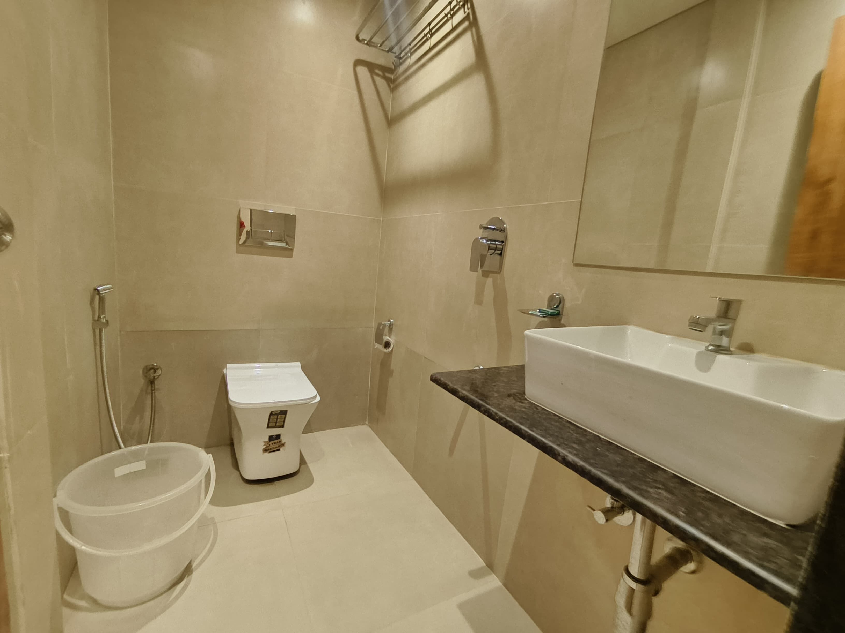 Un baño con lavamanos blanco, inodoro y ducha. Hay un espejo sobre el lavamanos y un perchero en la pared.
