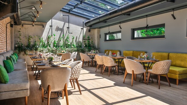 Un restaurante moderno con techo alto, tragaluces y plantas de interior. Las mesas están dispuestas con sillas y centros de mesa.