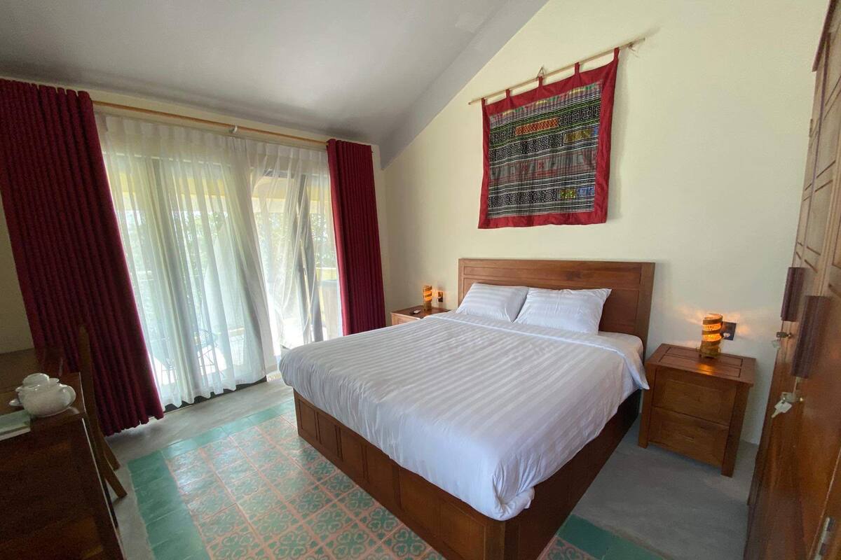 Une chambre à coucher bien aménagée, avec un grand lit, deux tables de chevet, une armoire en bois et une fenêtre ornée de rideaux légers.
