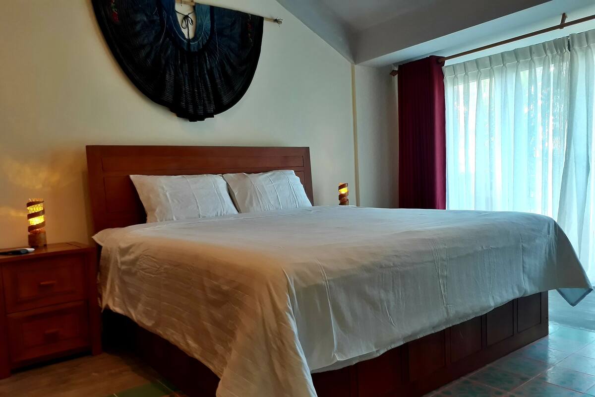 Une chambre d’hôtel bien éclairée, avec un grand lit, une table de chevet, une lampe fixée au mur et une fenêtre avec des rideaux.