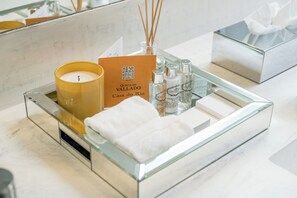 A glass tray on a bathroom counter with a candle, a tissue box, and several bottles of Castello gel. - Casa do Rio Wine Hotel - Vallado (Vila Nova de Foz Coa)