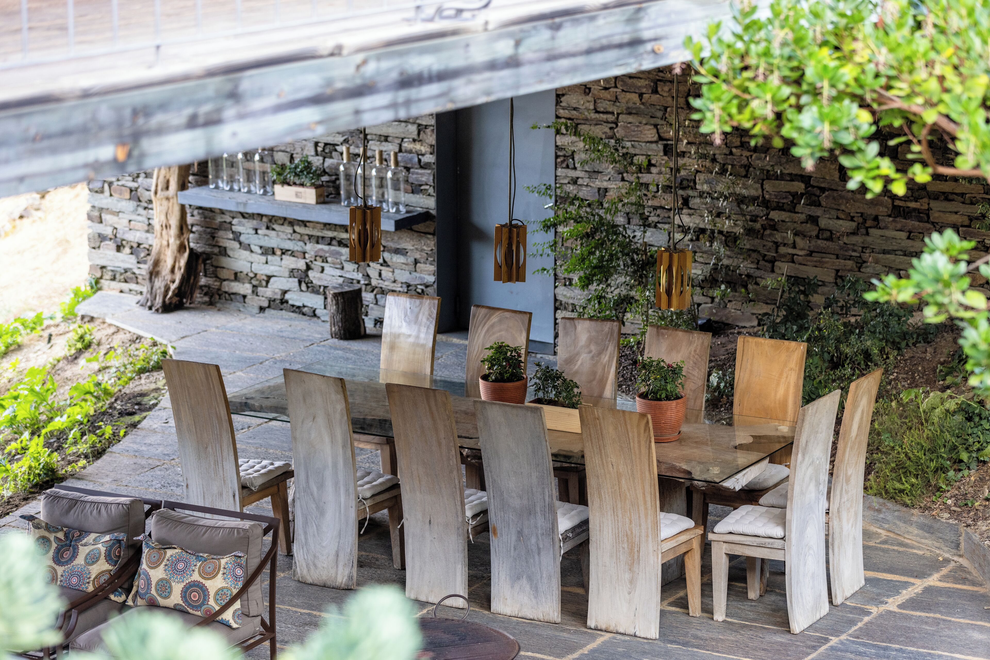 Un espacio de comedor al aire libre con sillas y mesas de madera, rodeado de plantas en macetas y un muro de piedra.