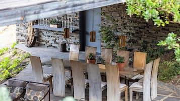 Un espacio de comedor al aire libre con sillas y mesas de madera, rodeado de plantas en macetas y un muro de piedra.