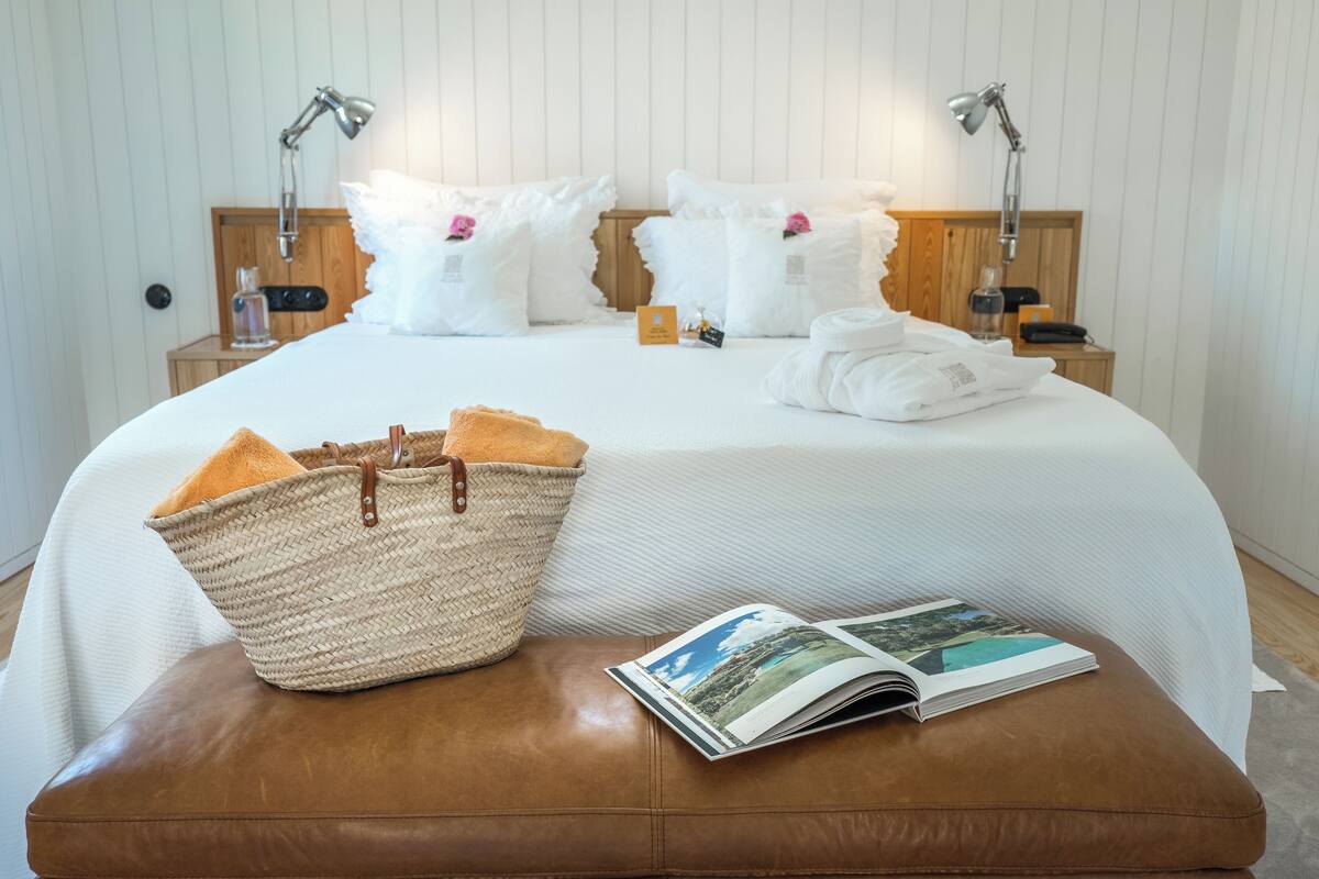 Una cama bien hecha con sábanas blancas, un reposapiés de cuero marrón, una cesta tejida y una revista sobre el reposapiés.