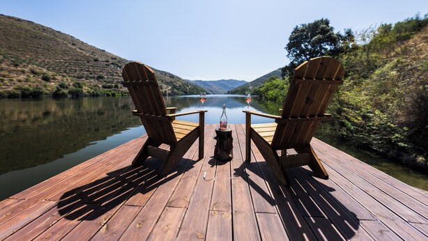 Dos sillas Adirondack mirando un lago tranquilo con una ladera boscosa al fondo.
