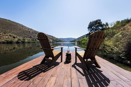 Two Adirondack chairs facing a calm lake with a forested hillside in the background.