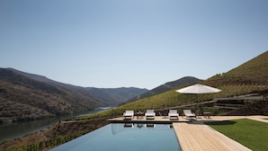 Zona junto a la piscina con sillones de descanso y una sombrilla, con vistas a un viñedo y un río.