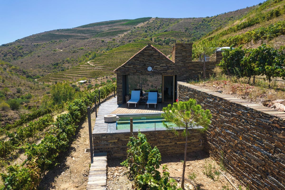 Edificio de piedra con piscina y sillones de descanso, rodeado de hileras de viñedos.