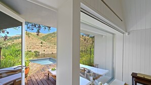 Una habitación moderna con terraza de madera, una mesa de vidrio y vistas a una piscina y colinas.