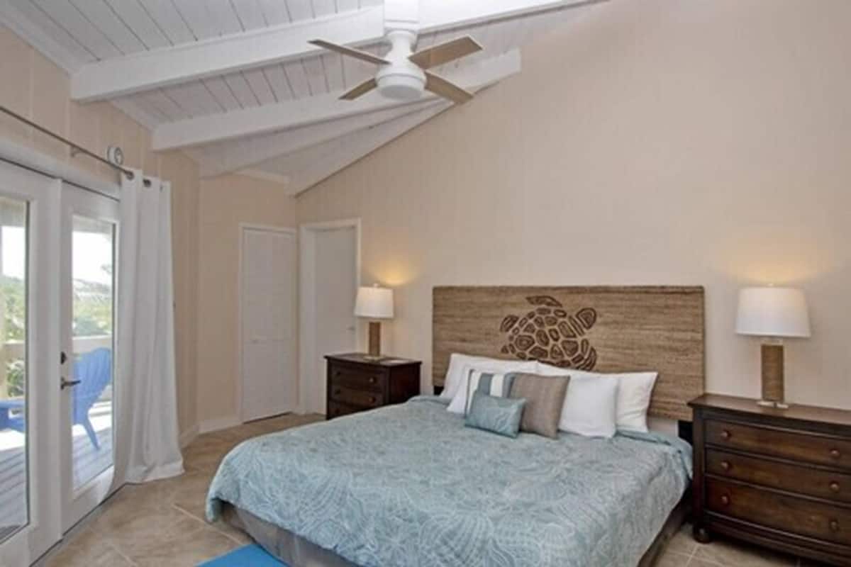 Un dormitorio con una cama, dos mesitas de noche, un ventilador de techo y un amplio ventanal con cortinas.
