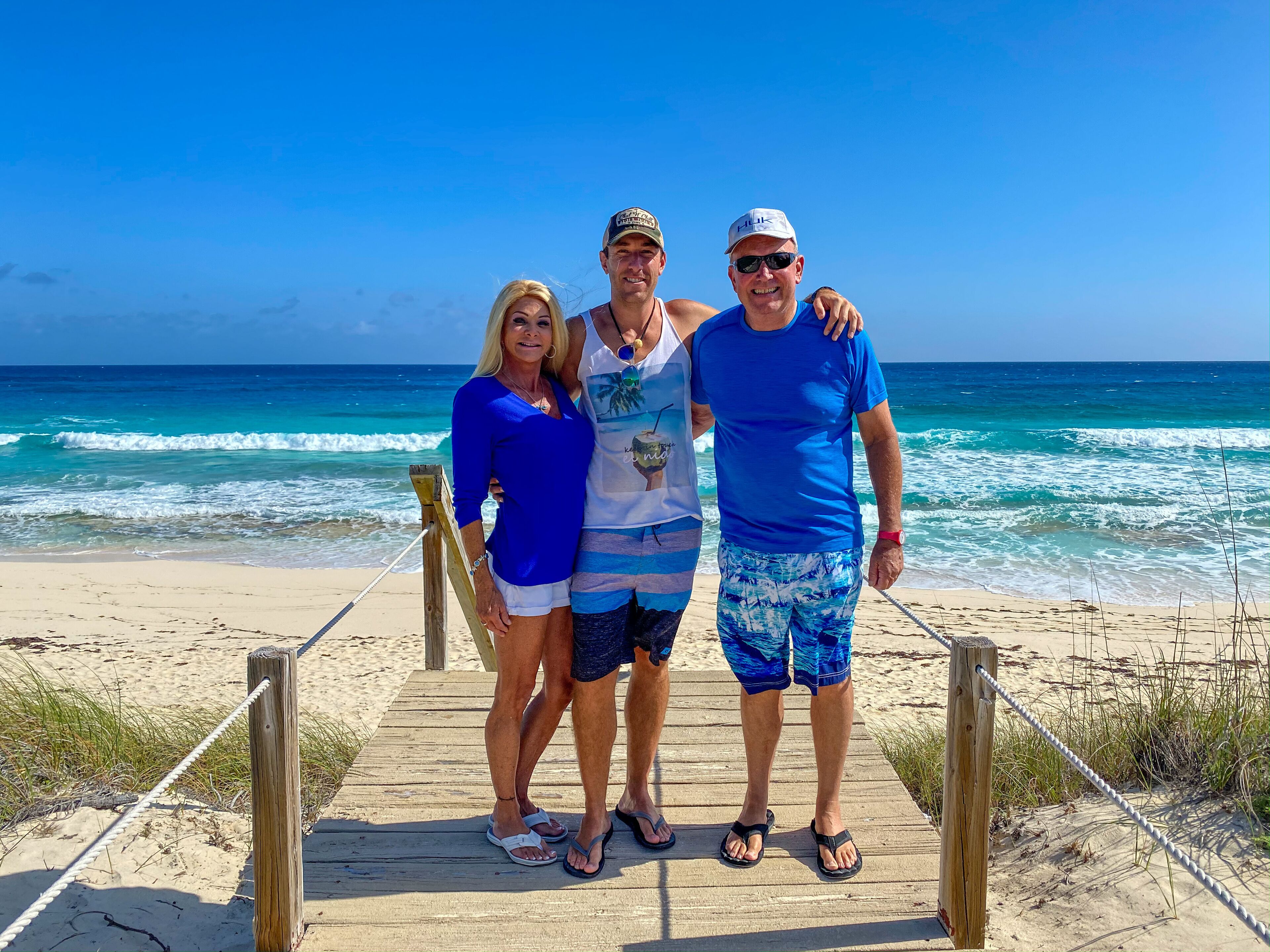 Three people standing on a wooden boardwalk at a beach with ocean waves in the background.