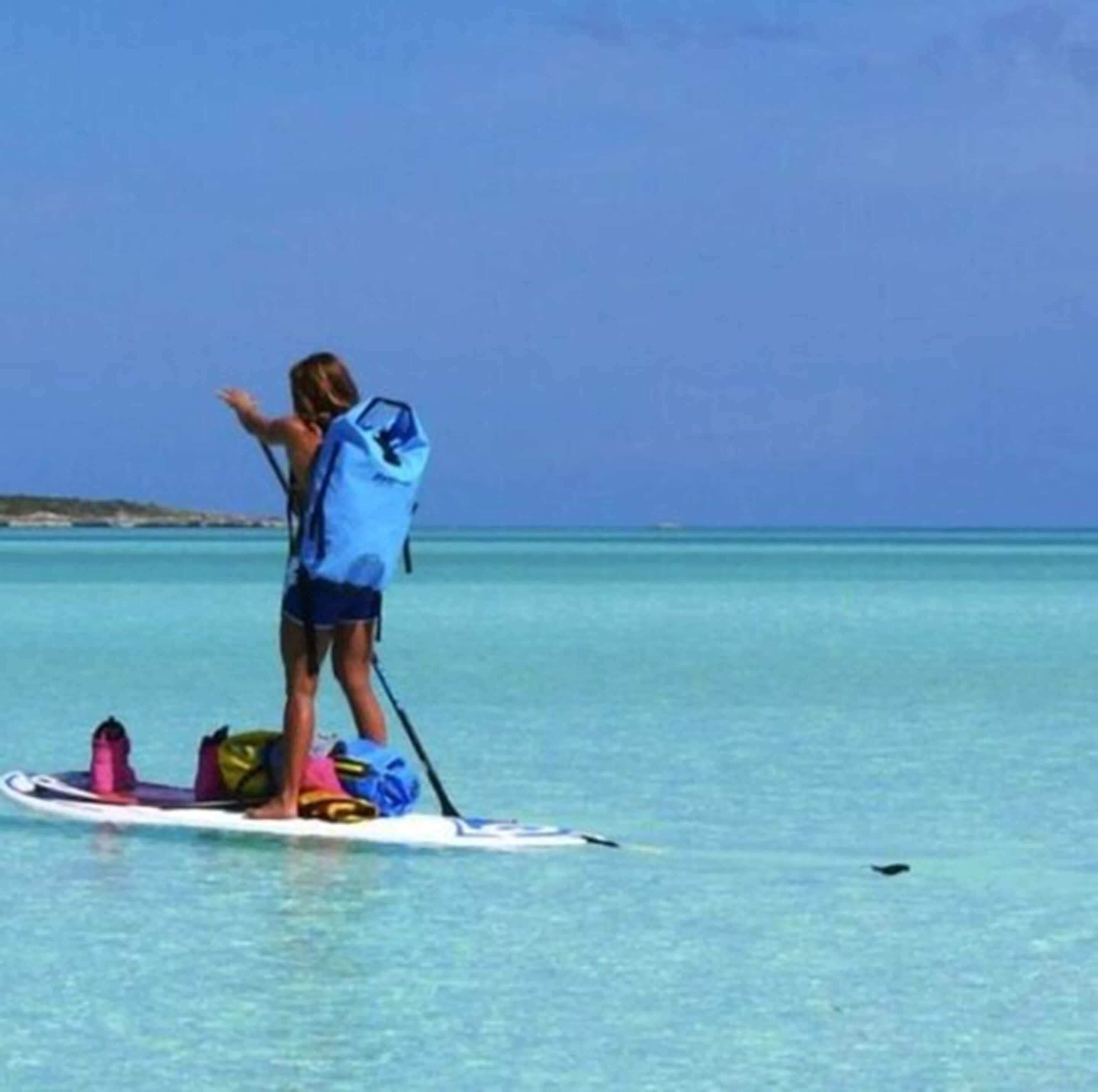 A person standing on a paddleboard in clear blue water, with a bag and paddle on the board.