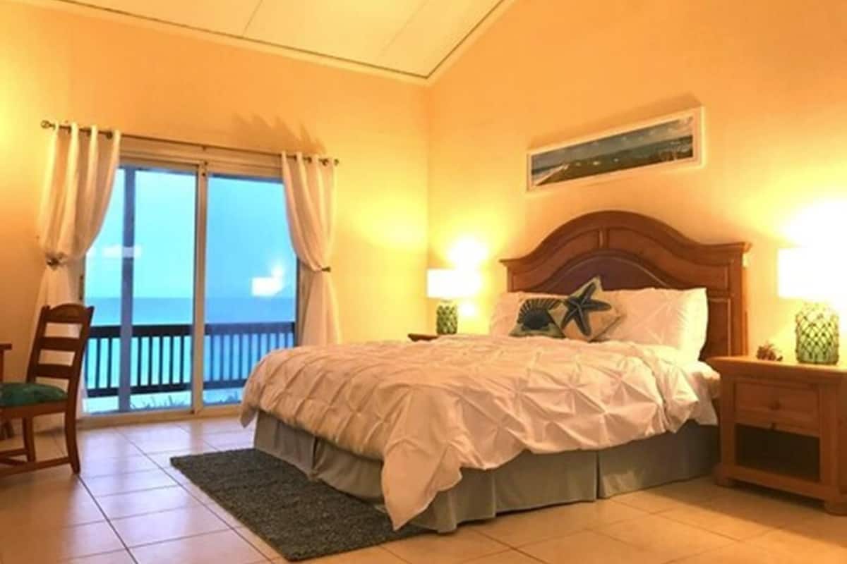 Un dormitorio con cama, mesita de noche, una silla, una ventana con cortinas y vista al océano.