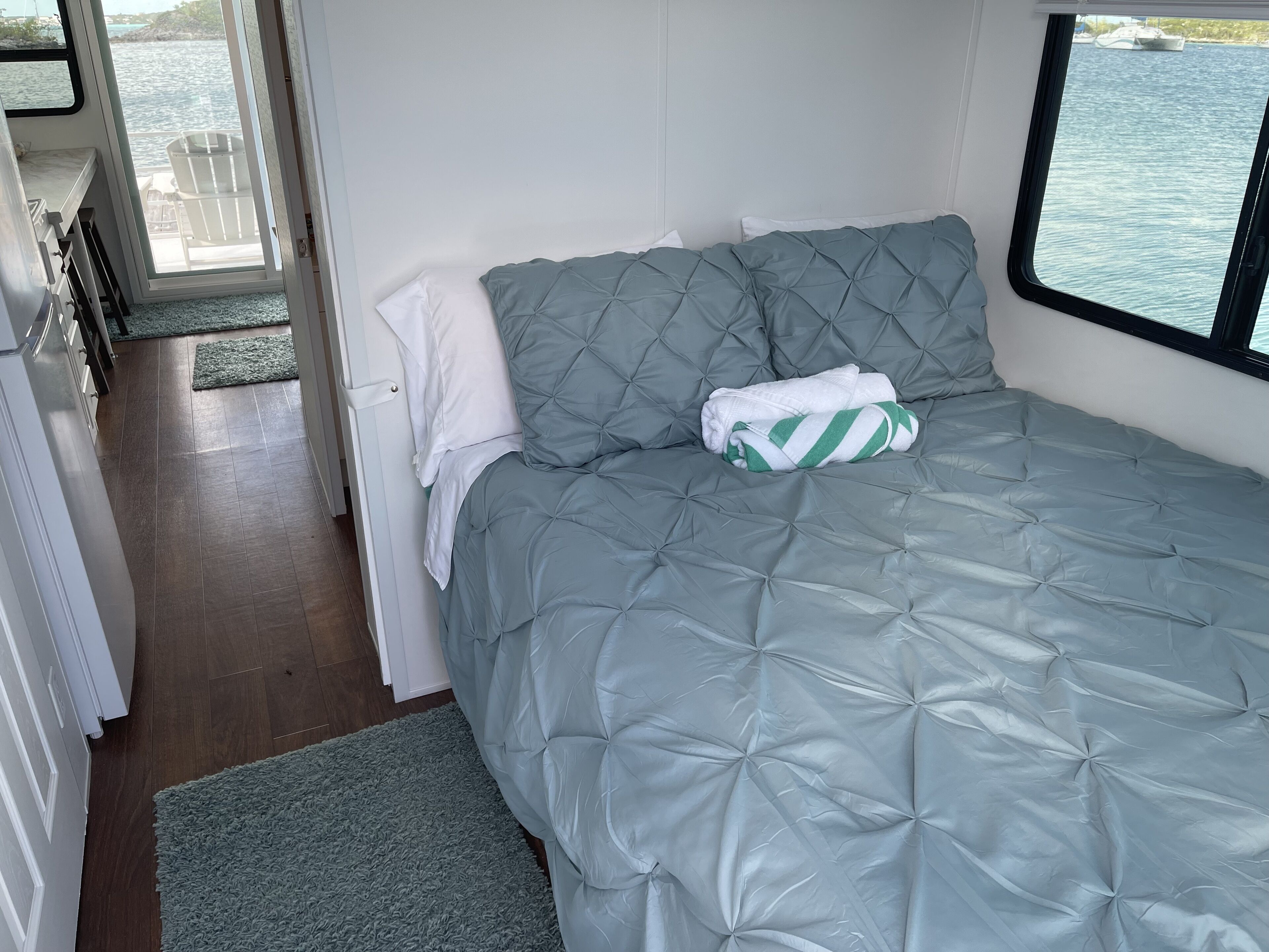 A compact bedroom with a grey quilted bedspread, a neatly folded white towel, and a green and white striped towel on the bed.