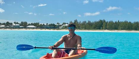 A person kayaking on clear turquoise water with a beach and palm trees in the background.