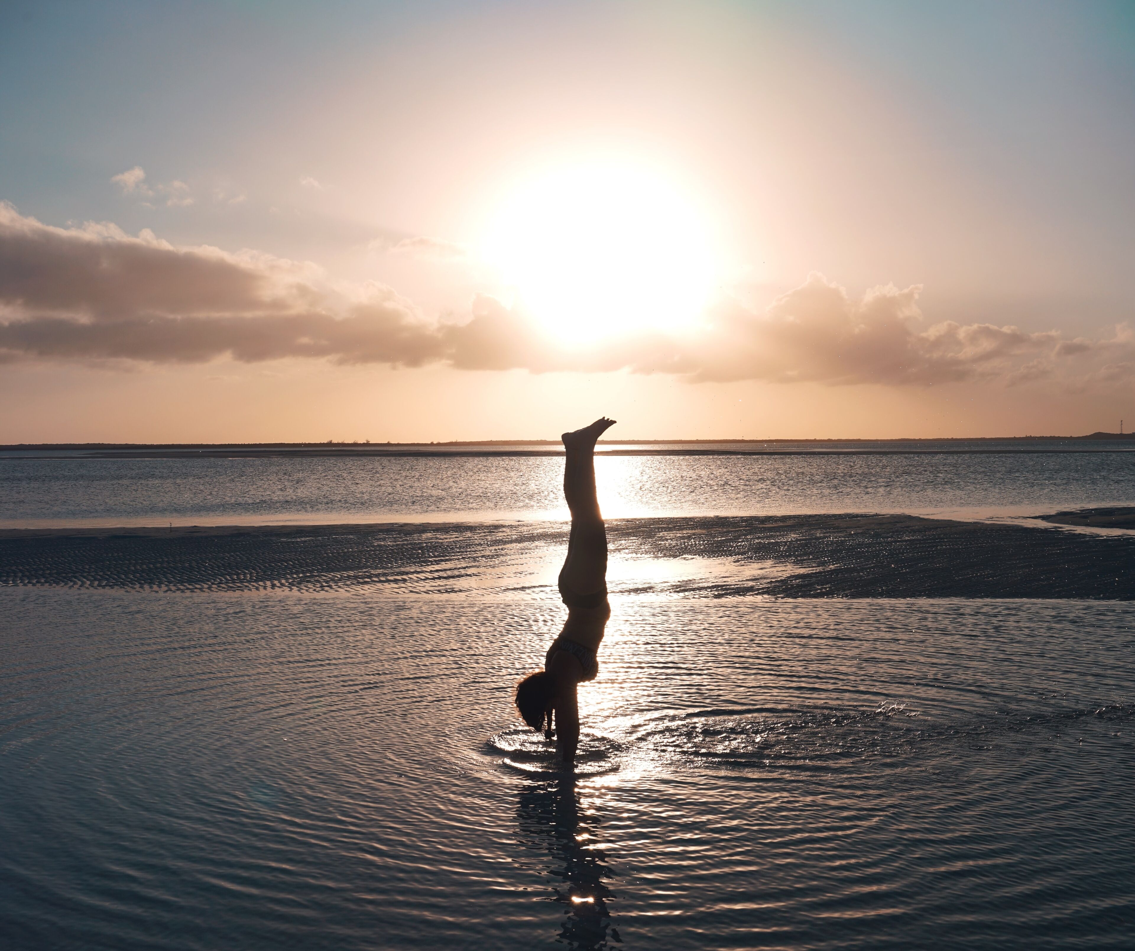 Eine Person macht eine Handstand in flachem Wasser, im Hintergrund ein Sonnenuntergang.