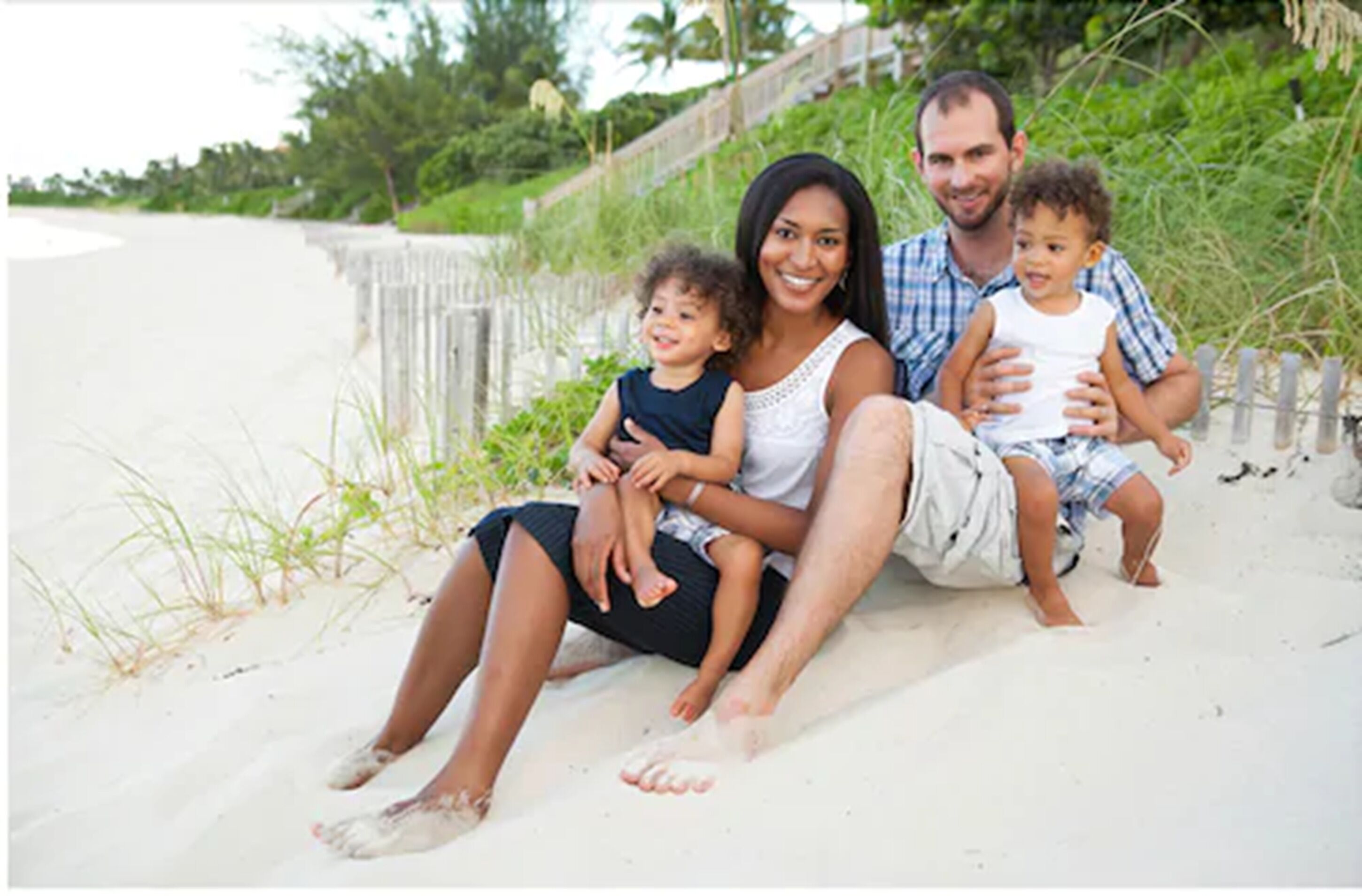 Una familia de tres en una playa arenosa con una valla de madera y vegetación al fondo.