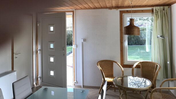 A room with a wooden ceiling, a glass table, wicker chairs, and a window with a view of greenery. - Kleines Ferienhaus (Berlin)