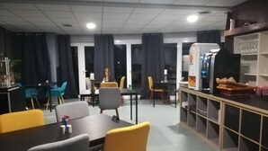 Un espace repas comprenant une longue table et des chaises, un réfrigérateur et une étagère où sont rangés des produits alimentaires.