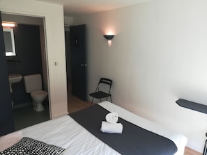 Habitación de hotel con una cama, una silla, una lámpara fijada a la pared y un baño visible a través de una puerta abierta.