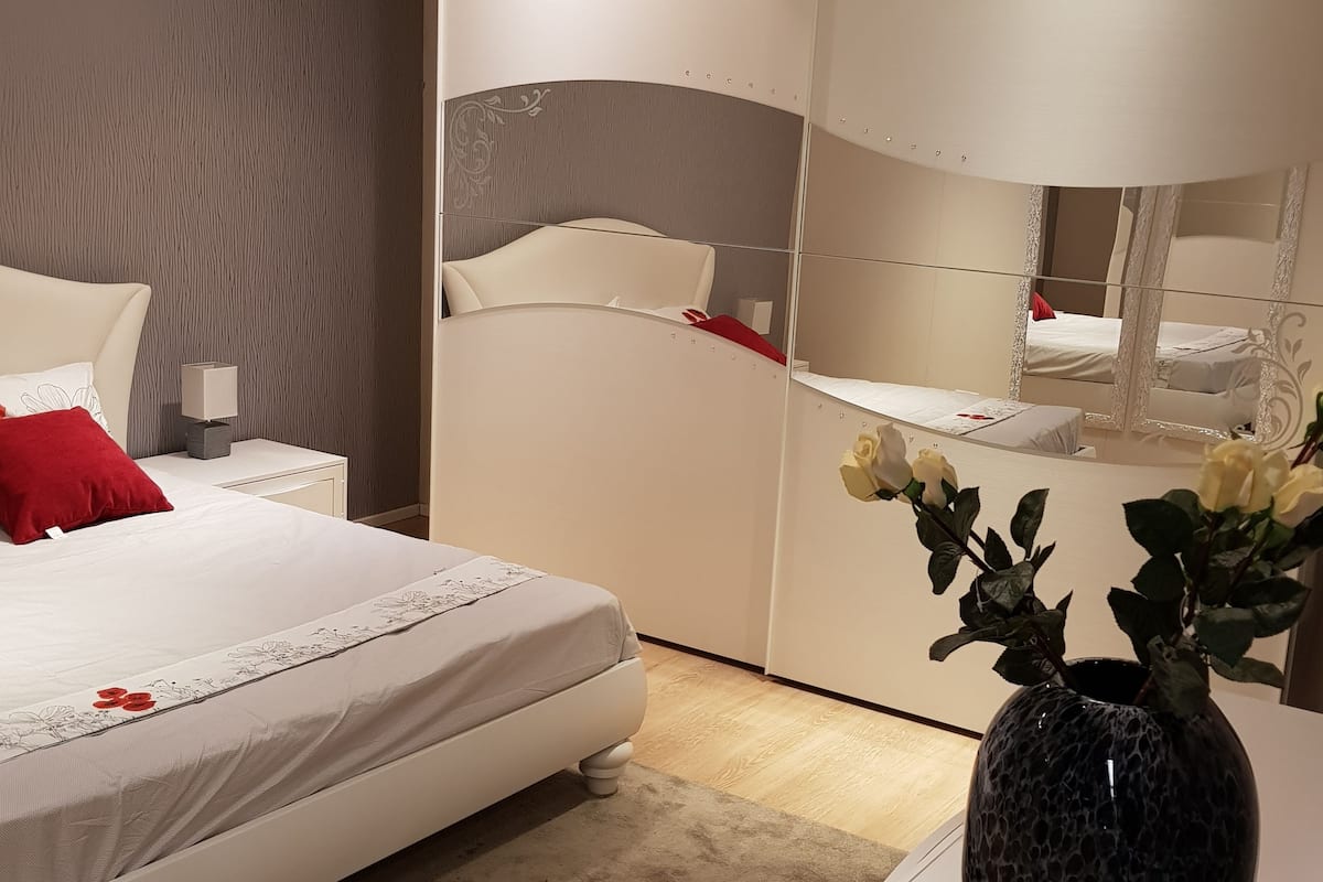A bedroom with a bed, a nightstand, a mirror, and a vase with flowers.