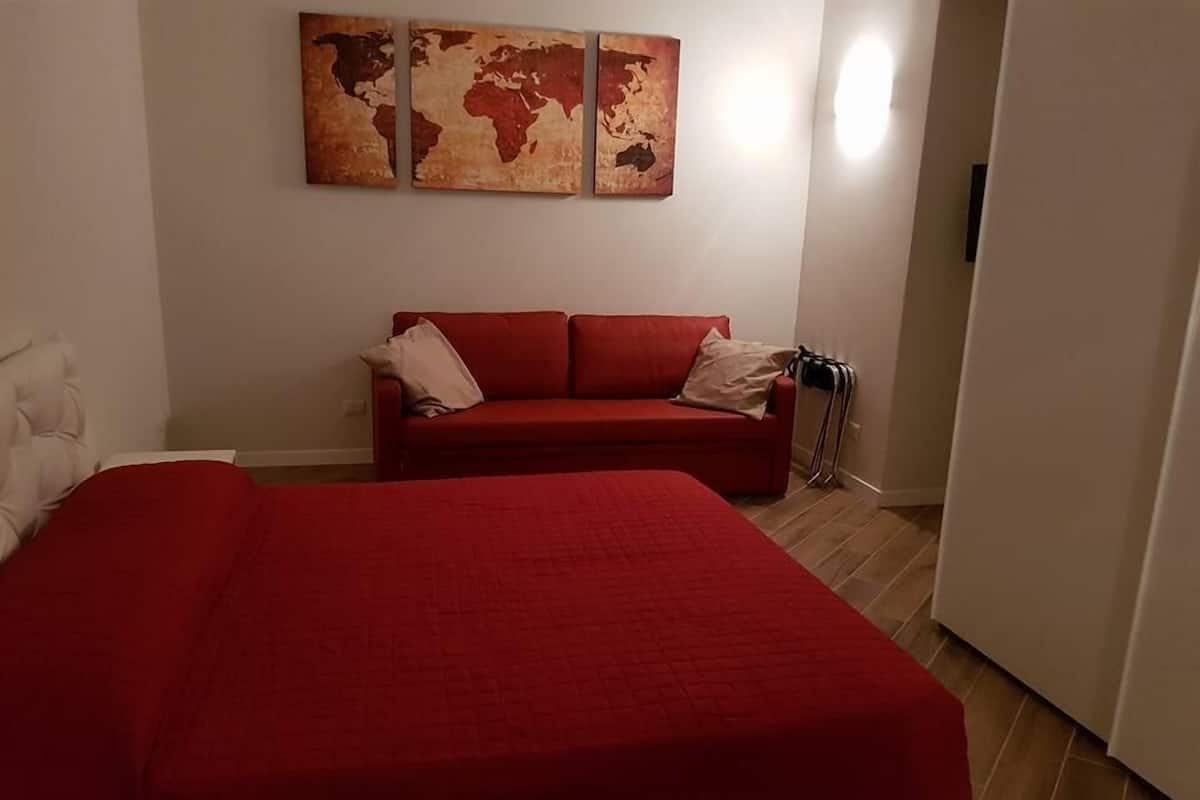 A room with a red bedspread, a red sofa, and a world map artwork on the wall.