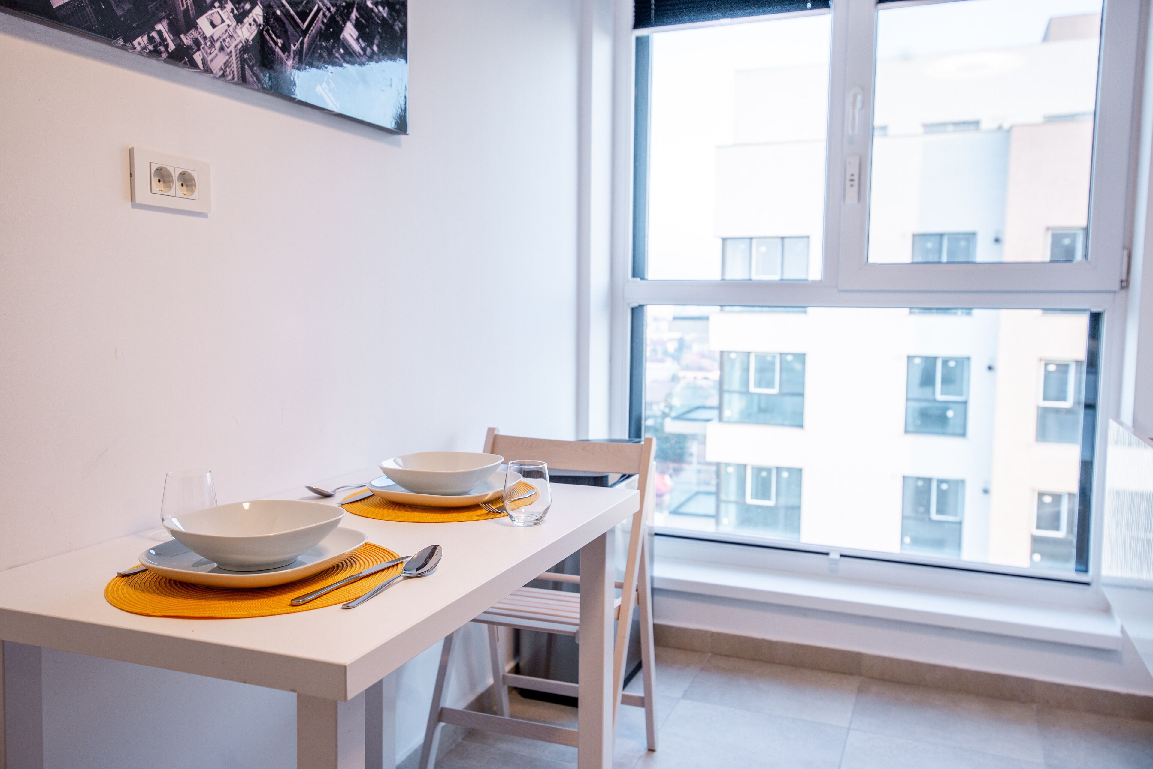 Un coin repas avec une table blanche dressée pour deux personnes, une fenêtre donnant sur des immeubles et un tableau accroché au mur.