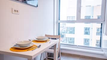 Un coin repas avec une table blanche dressée pour deux personnes, une fenêtre donnant sur des immeubles et un tableau accroché au mur.