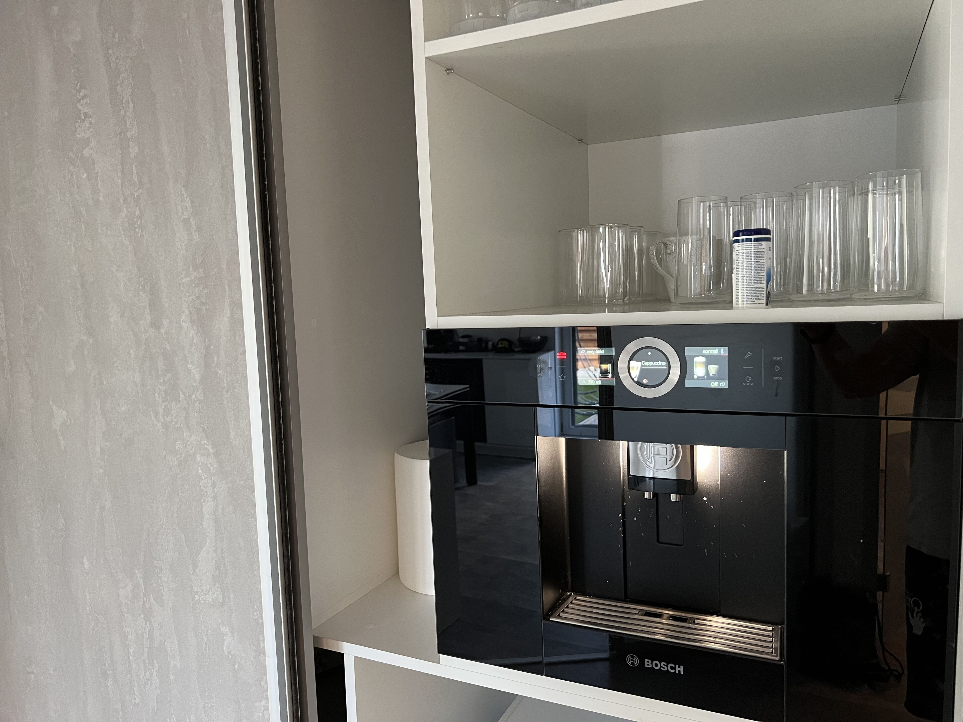 Une cuisine moderne équipée d’une machine à café Bosch, d’une étagère blanche où sont posés des verres transparents, et d’une fenêtre donnant sur l’extérieur.