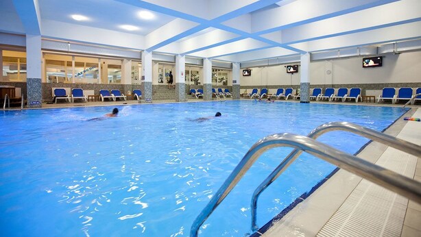 Una piscina interior con agua cristalina, una escalera de piscina y sillas de descanso azules.
